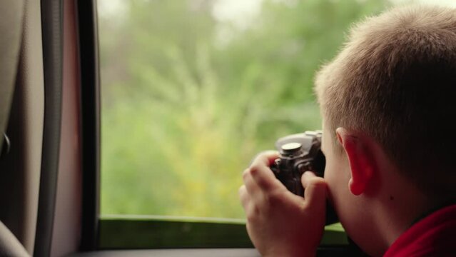 Child boy with camera is sitting in car, taking pictures of nature through window. Childrens journey. Family enjoy car travel on vacation. Child dreams of adventure, looks out open window of car