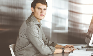 Young businessman working on his computer sitting at the desk in office. Headshot portrait of a man