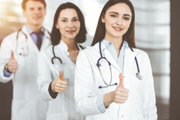 Group of young cheerful doctors standing as a team with thumbs up in sunny hospital. Medical help, insurance in health care, best disease treatment and medicine concept