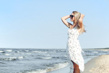 Fototapeta na wymiar Happy blonde beautiful woman on the ocean beach standing in a white summer dress and sun glasses, raising hands