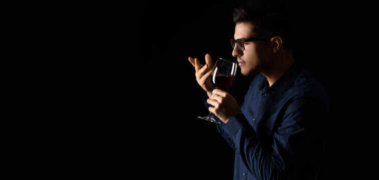 Young male sommelier tasting wine on dark background with space for text