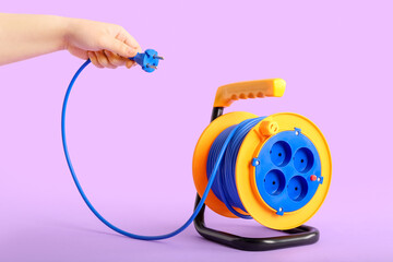 Female hand with extension electric cable reel on lilac background