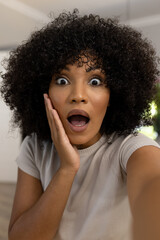 Portrait of surprised biracial woman with afro at home making video call, touching face in shock