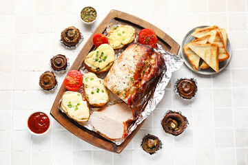 Wooden board with tasty baked meat, potatoes, tomatoes and mushrooms on light tile background
