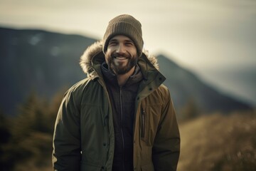 Fototapeta na wymiar Handsome bearded man in warm jacket and hat smiling at camera while standing on top of a mountain