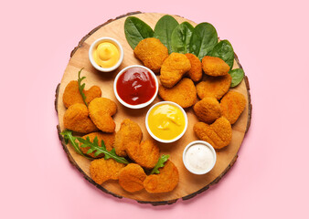 Wooden board with tasty nuggets and different sauces on pink background