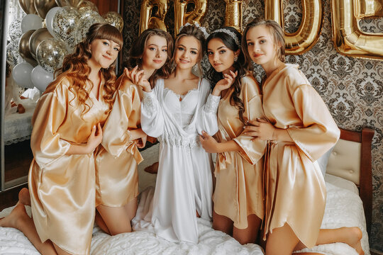 Young bridesmaids in silk robes in a hotel room. Beautiful women celebrating a bachelorette party sitting on the bed in themed pajamas.