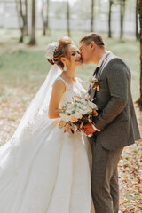 Fashionable groom and cute blonde bride in a white dress with a crown, a bouquet are hugging, laughing in the park, garden, forest outdoors. Wedding photography, portrait of smiling newlyweds.