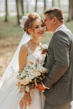 Fashionable groom and cute blonde bride in a white dress with a crown, a bouquet are hugging, laughing in the park, garden, forest outdoors. Wedding photography, portrait of smiling newlyweds.