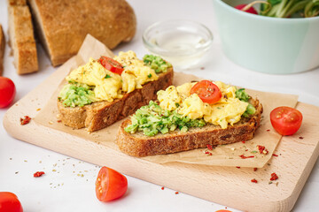 Wooden board with tasty scrambled eggs sandwich and vegetables on light background
