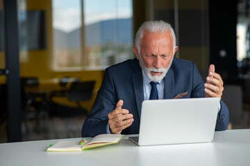 Handsome smiling senior man while sitting at his cozy workplace with laptop at home, retired male chatting with friends in social media,