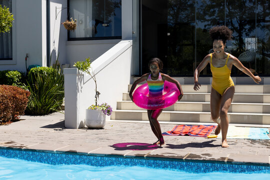 Happy biracial mother and daughter with inflatable jumping into swimming pool in sunny garden