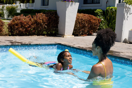 Happy biracial mother and daughter learning to swim with float in swimming pool in sunny garden