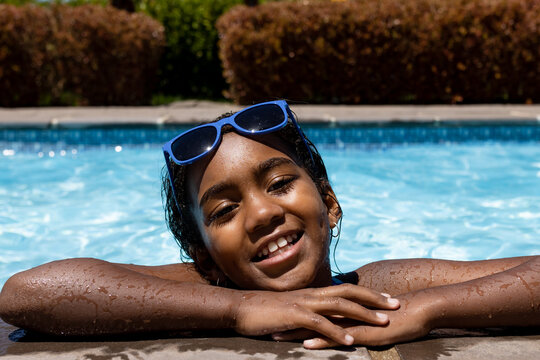 Portrait of happy biracial girl in water with sunglasses leaning on side of swimming pool