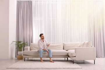 Happy man drinking coffee while resting on sofa near window with beautiful curtains in living room