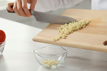 Professional chef putting cut garlic into bowl at white marble table indoors, closeup