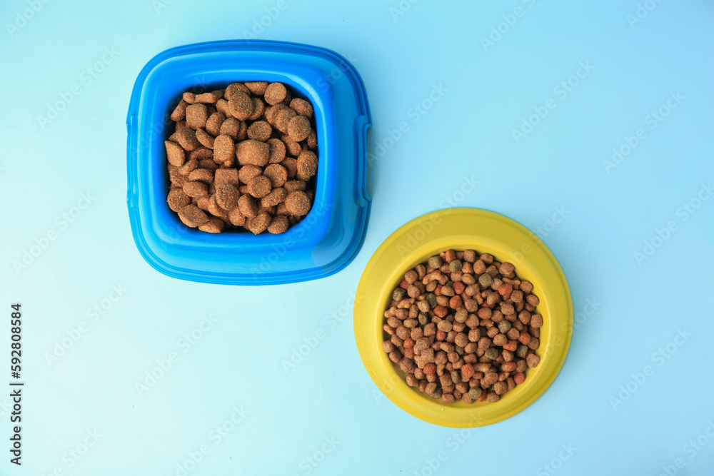 Wall mural Dry pet food in feeding bowls on light blue background, flat lay