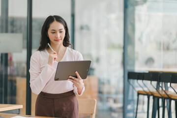 Fototapeta premium A portrait of a smiling, young, beautiful, professional, and confident millennial Asian businesswoman using a digital tablet to analyze sales data at a co-working space.