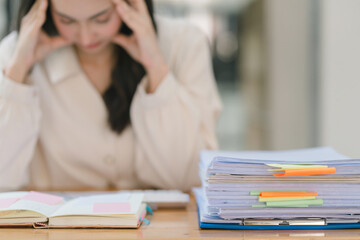 The stressed and exhausted millennial Asian businesswoman is seen sitting at her office desk with her hand on her head, indicating a hard working day where she is overloaded with work. Selective focus