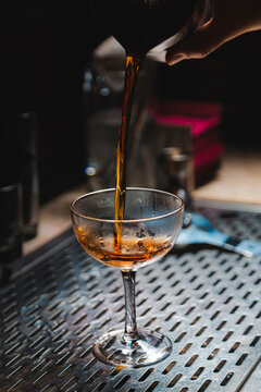 Manhattan style craft cocktail pouring into elegant coupe glass on bar top.