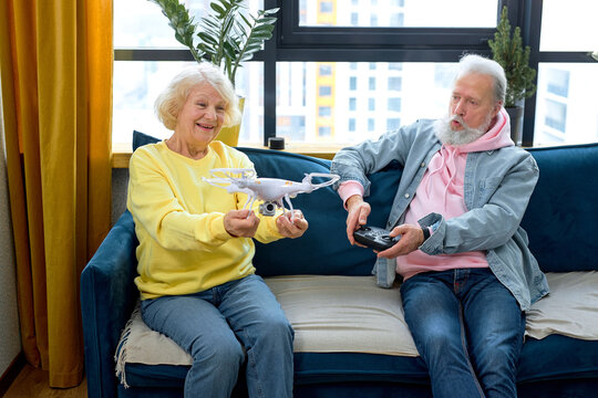 Happy elderly caucasian couple launch a drone at home, man holding console in hands. retired spouses in casual clothes enjoy using modern technologies, learning to use at home in living room