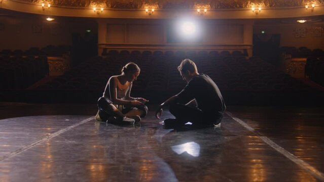 Couple of tired ballet dancers rest after choreography rehearsal. Ballerina sits with partner on theatre stage and discusses performance. Empty rows in theatrical hall. Concept of classical ballet art