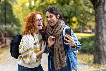 Two student woman friends in urban public park use mobile phone and smile