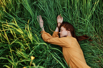 a close horizontal photo of a pleasant woman in a long orange dress resting lying in the tall grass with her eyes closed in sunny weather