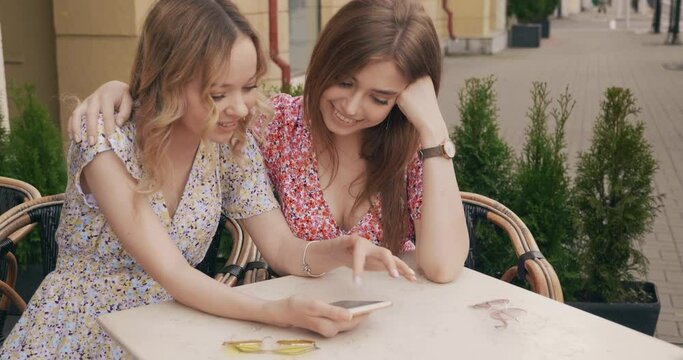 Two young beautiful smiling hipster girls in trendy summer sundress. Carefree women chatting in veranda cafe. Model shows her friend information in her smartphone. They watching photos after shopping 