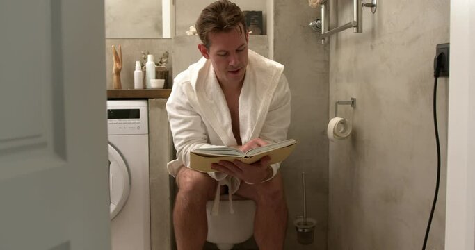 Male reading book while sitting on toilet bowl at home, relaxing, having rest. Young caucasian guy in domestic bathrobe in the morning, in broght cozy bathroom. lifestyle, people, everyday life