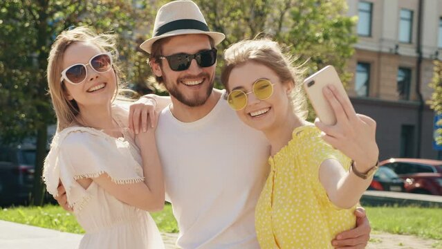 Group of young three stylish friends posing in the street. Fashion man and two cute girls dressed in casual summer clothes.  Cheerful women taking selfie photos