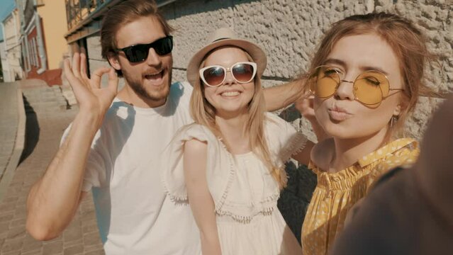 Group of young three stylish friends posing in the street. Fashion man and two cute girls dressed in casual summer clothes. Smiling models looking at camera. Cheerful women taking selfie photos