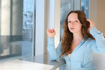 young woman straightening her hair waiting for friends at the window in cafe at school in a...