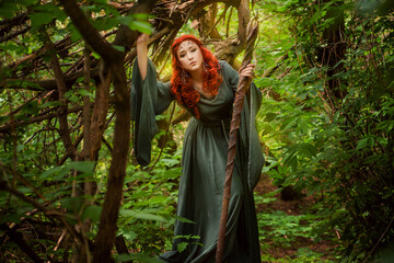 A young woman in a cloak, with a staff and a green dress in the form of a druid. Forest witch with...