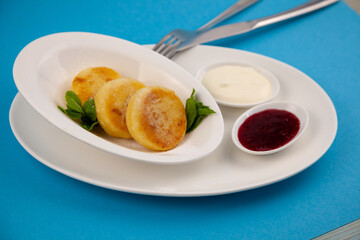 Round cottage cheese pancakes in a white plate on a blue background. Food breakfast. Healthy food. Sour cream sauce and mint. Top view closeup. Dessert meal. Curd cheese pancake ricotta fritters.