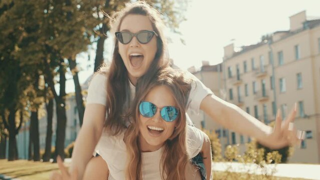 
Two young beautiful smiling hipster girls in trendy summer white t-shirt clothes.Sexy carefree women posing on street background. Model jumping on her friend back, gives piggyback riding outdoors