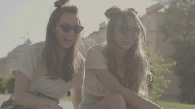 Not coloured video. Two young beautiful smiling hipster girls in trendy summer clothes. Sexy carefree women sitting on the stairs on street background. Positive models communicating in sunglasses