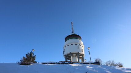 An observation tower in Mikkeli. The white tower on top of hill which is photographed from below. Clear blue sky and snow is in a picture. Three street lamps are near the tower. A sunny winter day.