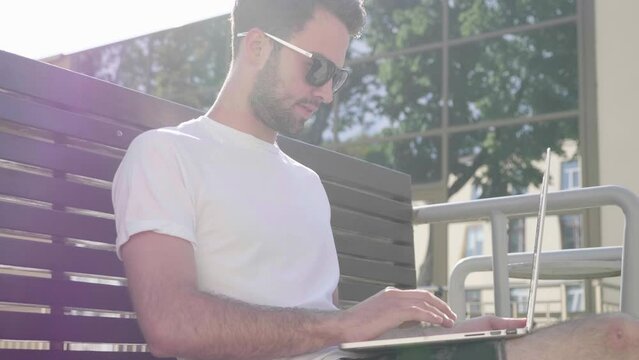 Handsome smiling stylish hipster lambersexual model. Fashion man sitting on the bench in the street and using laptop computer, writing text. Concept of co-working startup. Technology and communication