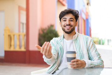 Handsome Arab man holding a take away coffee at outdoors inviting to come with hand. Happy that you came