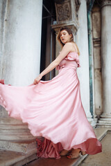 Fototapeta premium Fashion portrait of a stylish young woman with wavy blond hair in an elegant pink dress. The model poses in a beautiful dress against the backdrop of classical architecture. Fashion and style concept.