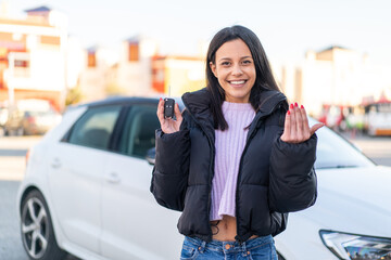 Young woman at outdoors holding car keys at outdoors inviting to come with hand. Happy that you came