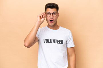 Fototapeta na wymiar Young volunteer caucasian man isolated on beige background with glasses and surprised