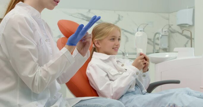 blonde brave girl is not afraid of dentist, she hold mirror, looks at it, female red-haired doctor putting ob blue gloves, preparing to pull a tooth. teeth care Orthodontic examination