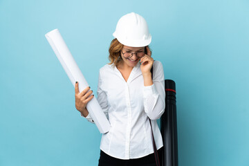 Young architect Georgian woman with helmet and holding blueprints over isolated background laughing