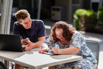 Happy young disability worker and his client with down syndrome working on devices at table