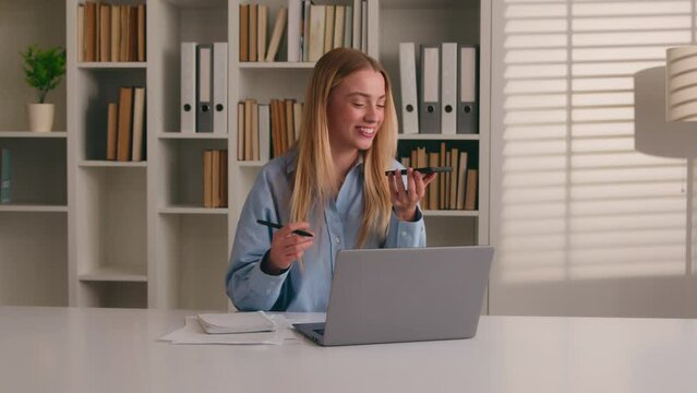 Multitasking Caucasian woman working with laptop writing in notebook talking mobile phone call loudspeaker happy businesswoman girl write notes typing computer record audio voice mail message studying