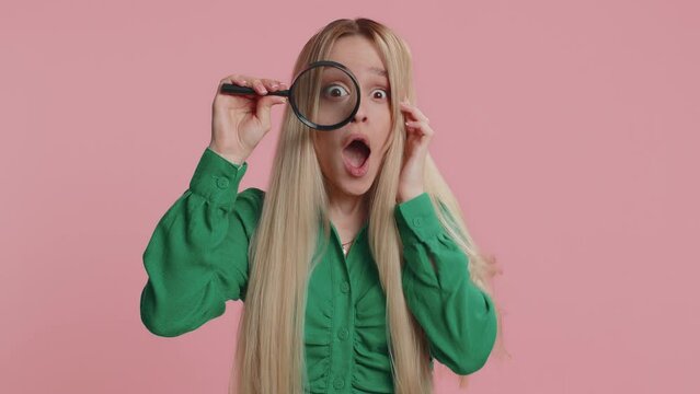 Investigator researcher scientist woman holding magnifying glass near face looking into camera with big zoomed funny eyes, searching analysing. Excited teen girl isolated on pink studio background