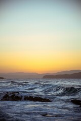 Vertical shot of sea waves on the shore with mountain silhouettes on the horizon during sunset