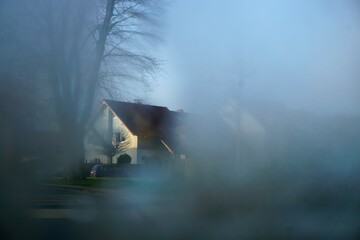 Bäume auf Wiese vor Einfamilienhäusern und Himmel bei Nebel und Sonne am Morgen Frühling